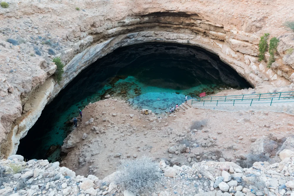 Bimmah Sinkhole with turquoise water inside a large limestone cavern, surrounded by rocky terrain and a stairway leading down to the pool.