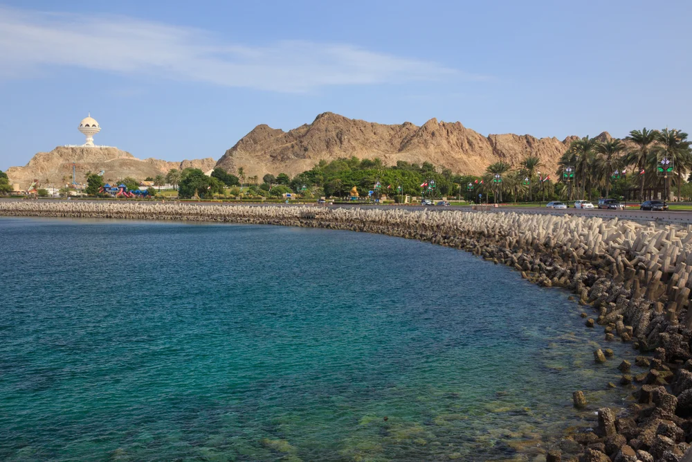 Scenic view of Muscat, Oman, with turquoise sea, rocky mountains, palm trees, and a waterfront breakwater